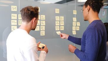 2 people brainstorm their campaigns with sticky notes on glass manifestations in the office